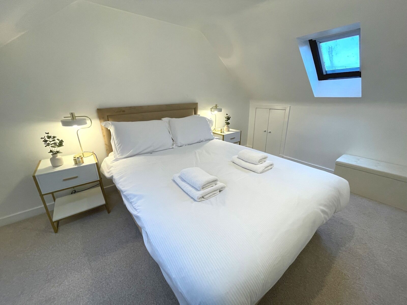 Een keurig opgemaakt slaapkamer met een groot bed, nachtkastjes met lampen, een klein raam en een kledingkast.