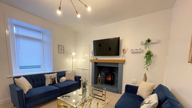 A living room with two blue sofas, a glass coffee table, a fireplace, and a mounted TV. - Beautifully Presented, Cosy Town Centre Cottage - Pass the Keys (Dumfries)