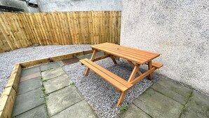 A wooden picnic table in a garden area with a stone pathway and a wooden fence in the background. - Beautifully Presented, Cosy Town Centre Cottage - Pass the Keys (Dumfries)