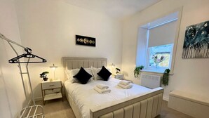 A neatly arranged bedroom with a bed, bedside tables, a radiator, a window with a view, and a wall-mounted artwork. - Beautifully Presented, Cosy Town Centre Cottage - Pass the Keys (Dumfries)