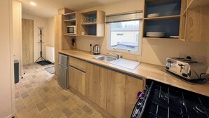 A compact kitchen with wooden cabinets, a built-in microwave, a toaster, and a window providing natural light. - Cosy 3BR Getaway in Beautiful Country Park - Pass the Keys (Beattock)