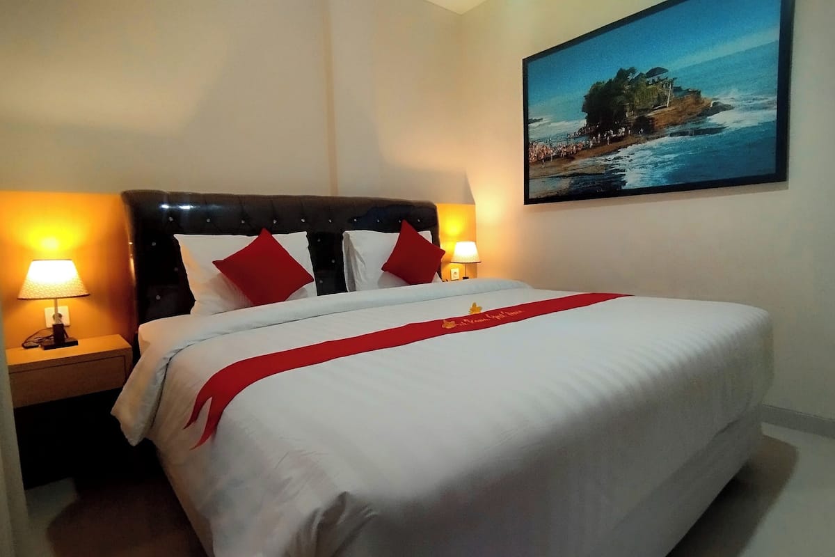 Una cama bien hecha con ropa blanca y detalles rojos, dos lámparas de noche y un cuadro de una escena costera en la pared.