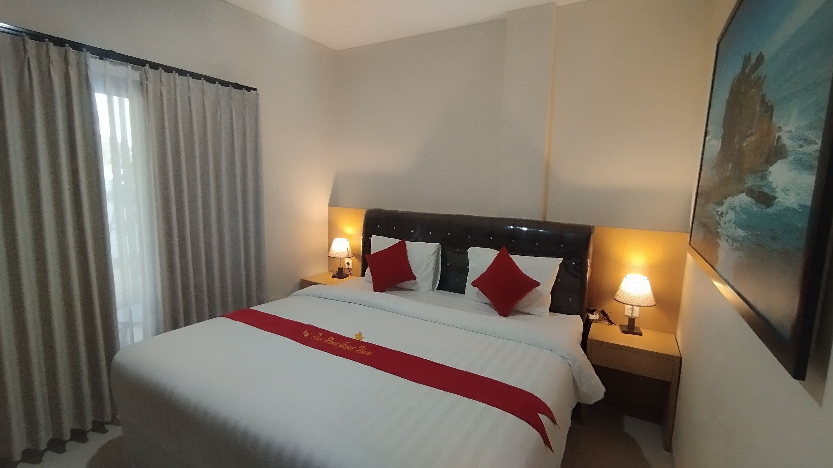 A hotel room with a large bed, two bedside lamps, a red throw pillow, and a painting on the wall.