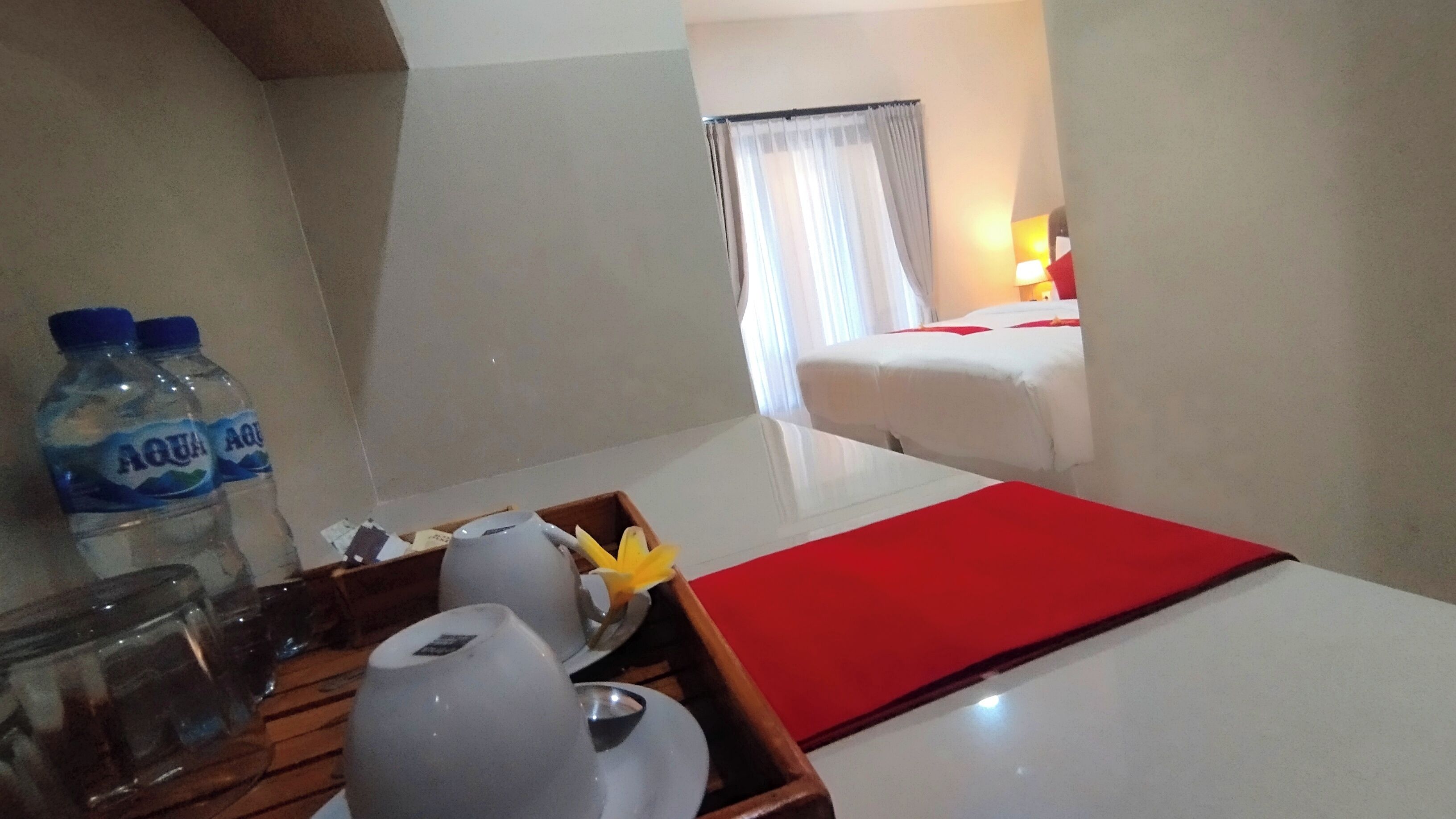 A hotel room with a bed, a nightstand with a lamp, a tray with a water bottle, and a red table runner.