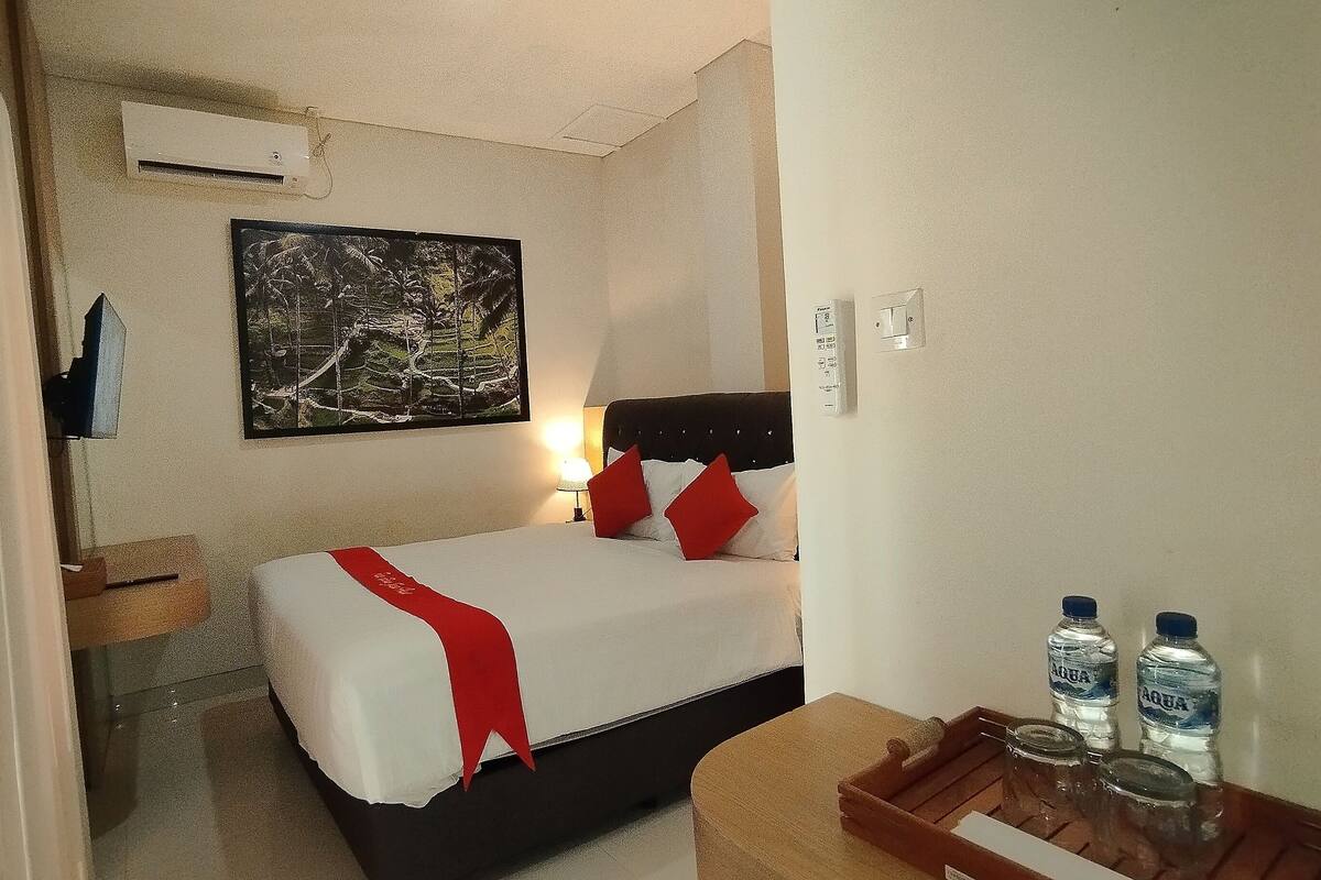 Un dormitorio con una cama, un mesita de noche, una lámpara, un aire acondicionado instalado en la pared y un cuadro.