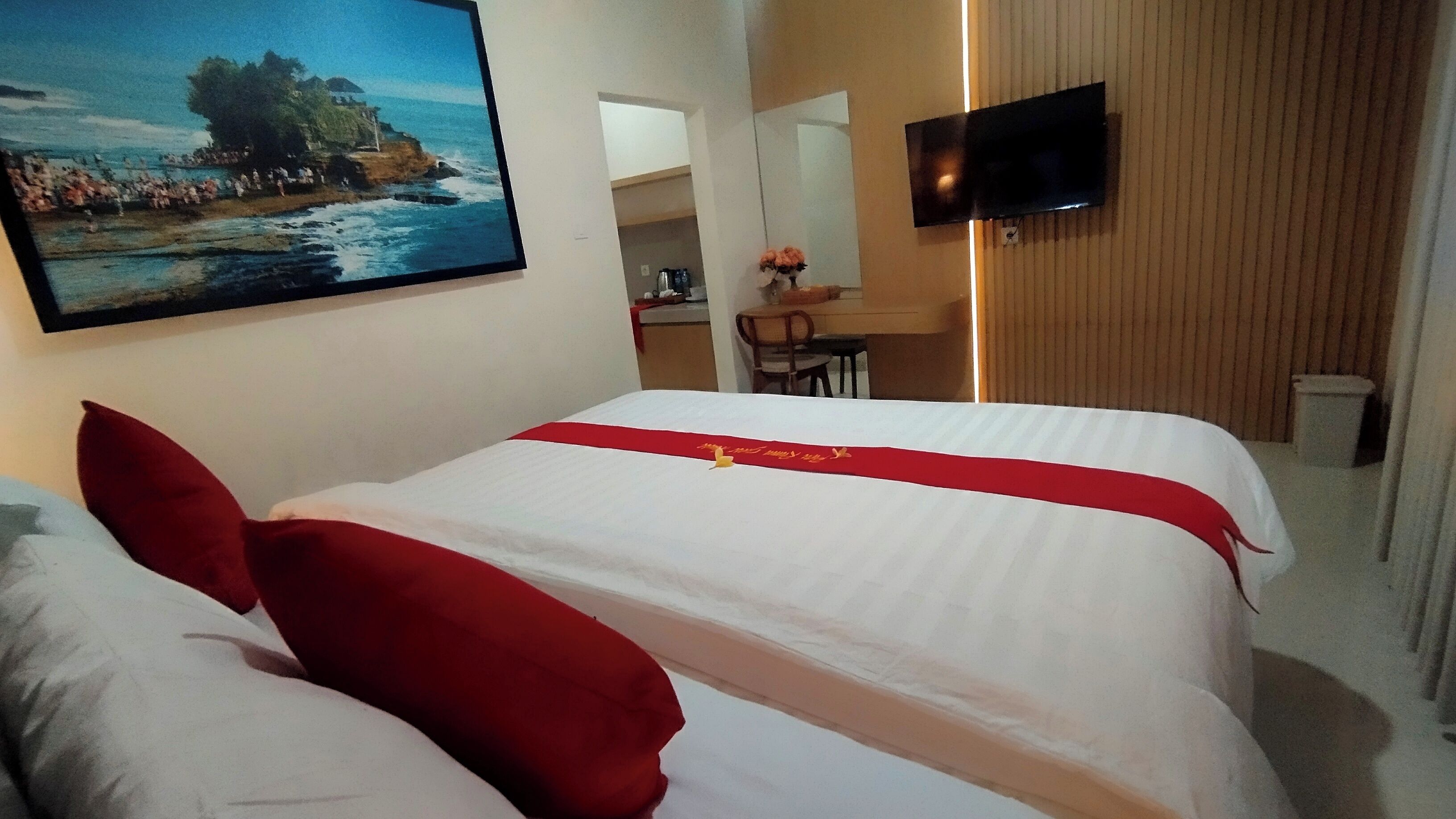 A hotel room with a bed, pillows, a red bedspread, a painting of a coastal scene, a TV, and a wooden wall.