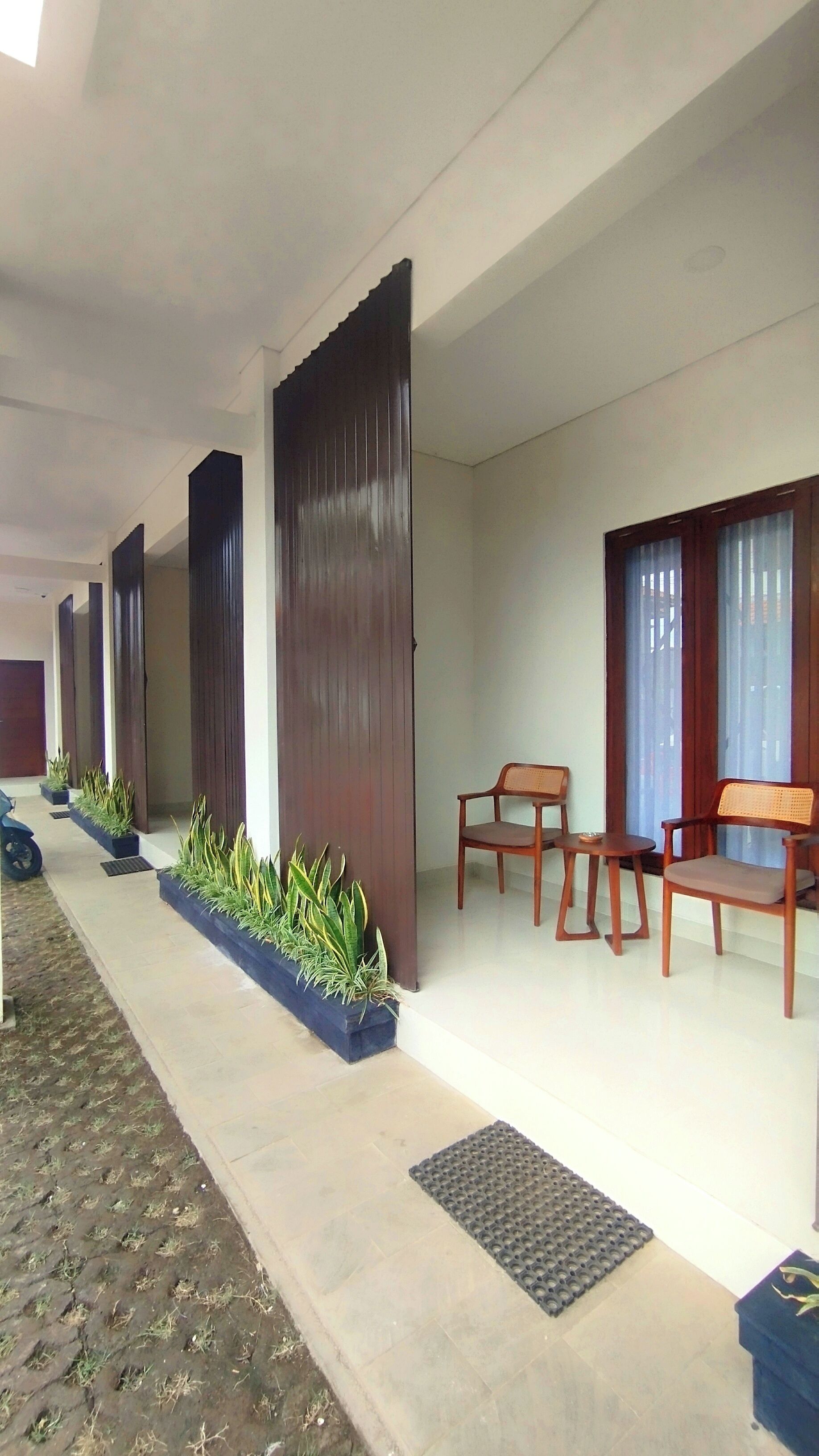 A covered outdoor area with a row of wooden chairs and tables, potted plants, and a tiled floor.