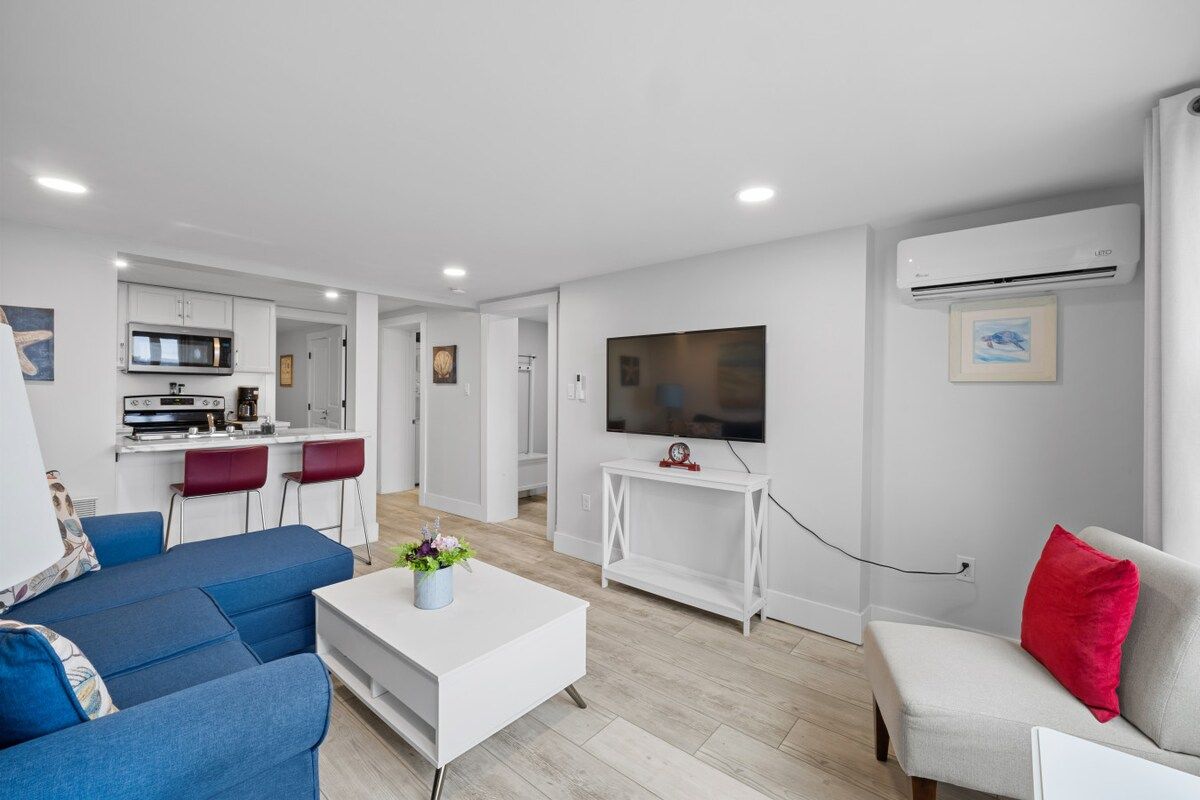 En moderne stue med sofa, sofabord, tv og et køkken i baggrunden.