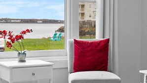 Ein Zimmer mit Blick auf einen Strand und ein Gebäude gegenüber dem Wasser. Ein weißer Tisch mit Pflanze und ein rotes Kissen auf einem grauen Stuhl.