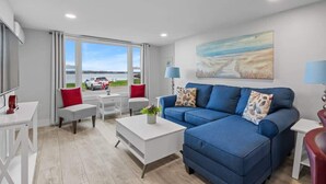 Ein modernes Wohnzimmer mit einem blauen Sofa, einem weißen Couchtisch und roten sowie weißen Sesseln. Ein großes Fenster bietet Blick auf ein Auto und die Landschaft.
