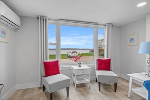 A room with a large window, a white air conditioning unit, a small table with a vase, two grey chairs with red cushions, and a blue chair. - Waterfront Deluxe 1 Bedroom Condo Downtown Ch'town (Charlottetown)
