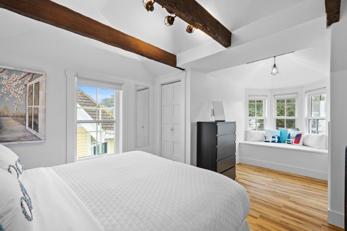 Une chambre à coucher avec un grand lit, une commode, un coin fenêtre avec des coussins et un tableau au mur.