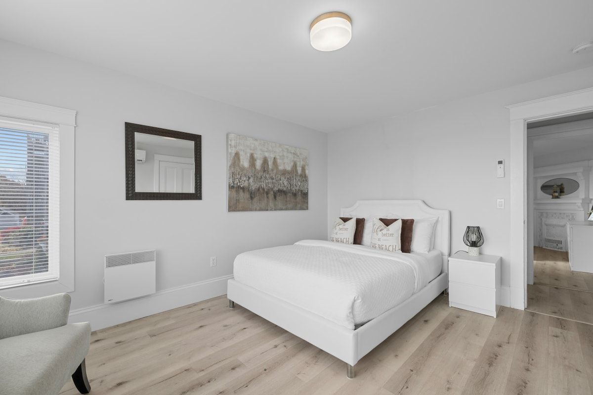 Un dormitorio con cama blanca, mesitas de noche, radiador, ventana con persianas y un cuadro en la pared.