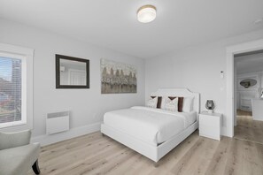Ein Schlafzimmer mit einem weißen Bett, Nachttischen, einem Heizkörper, einem Fenster mit Jalousien und einem Bild an der Wand.