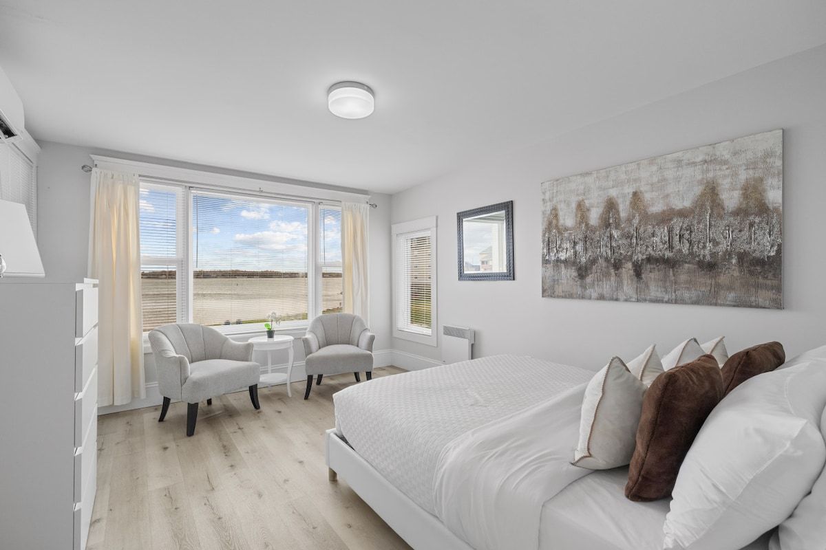 Une chambre à coucher avec un grand lit, deux chaises, une petite table et une fenêtre donnant sur un beau paysage.