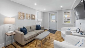 Un salon moderne avec un canapé beige, un fauteuil blanc et une table basse en bois. On y trouve des œuvres d’art murales sur le thème de la marine et un téléviseur à écran plat fixé au mur.