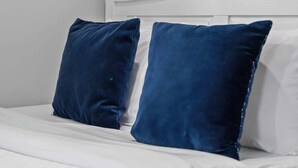 Un lit avec une literie blanche et deux oreillers bleu foncé, une tête de lit blanche et un élément décoratif en bois fixé au mur.