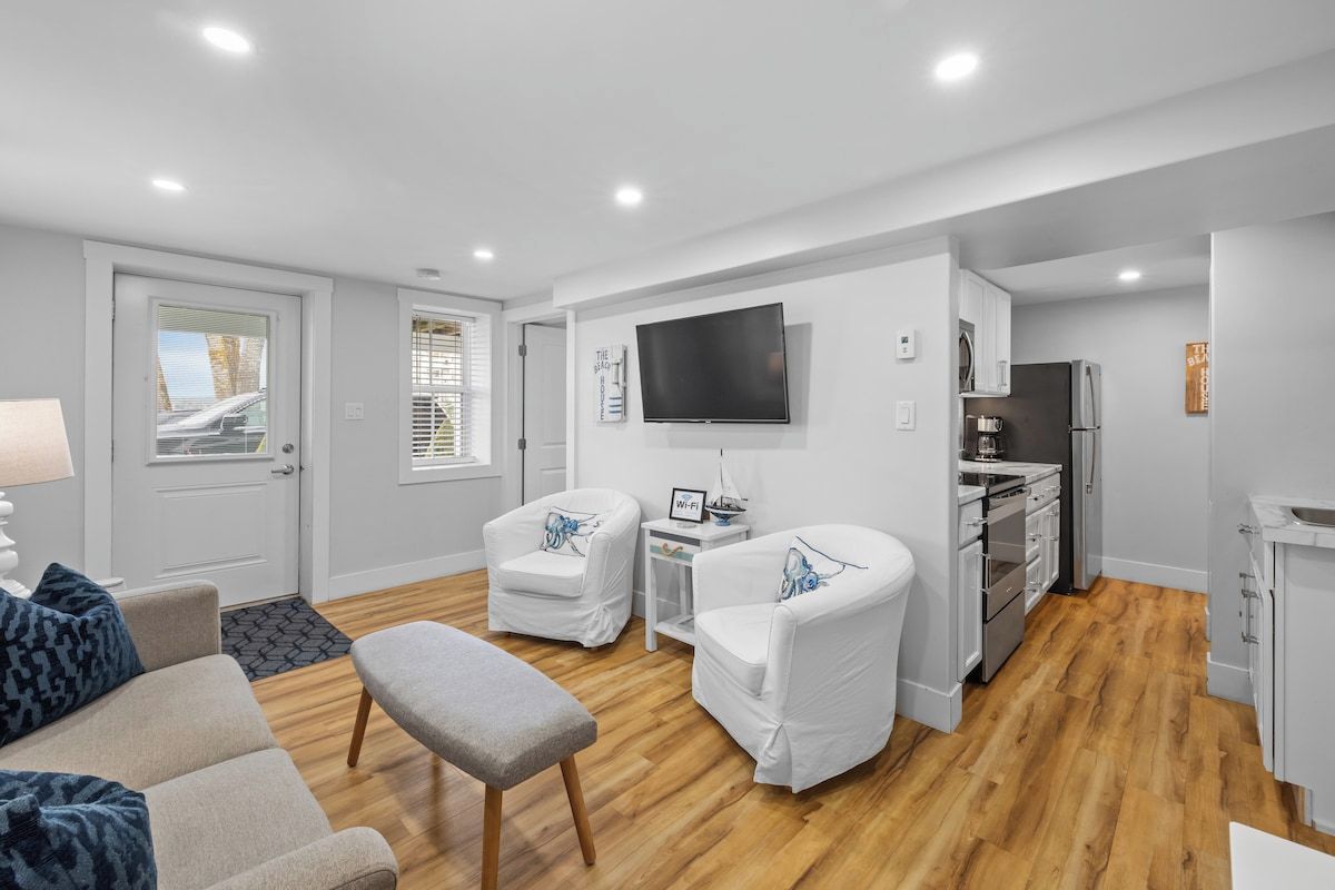 A modern living room with a sofa, two white armchairs, a coffee table, and a flat-screen TV mounted on the wall.