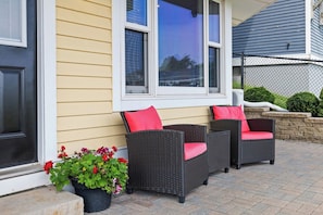 A house exterior with a porch, two wicker chairs with red cushions, and a potted plant with red flowers. - Waterfront Posh King Studio Suite Downtown Ch'town (Charlottetown)