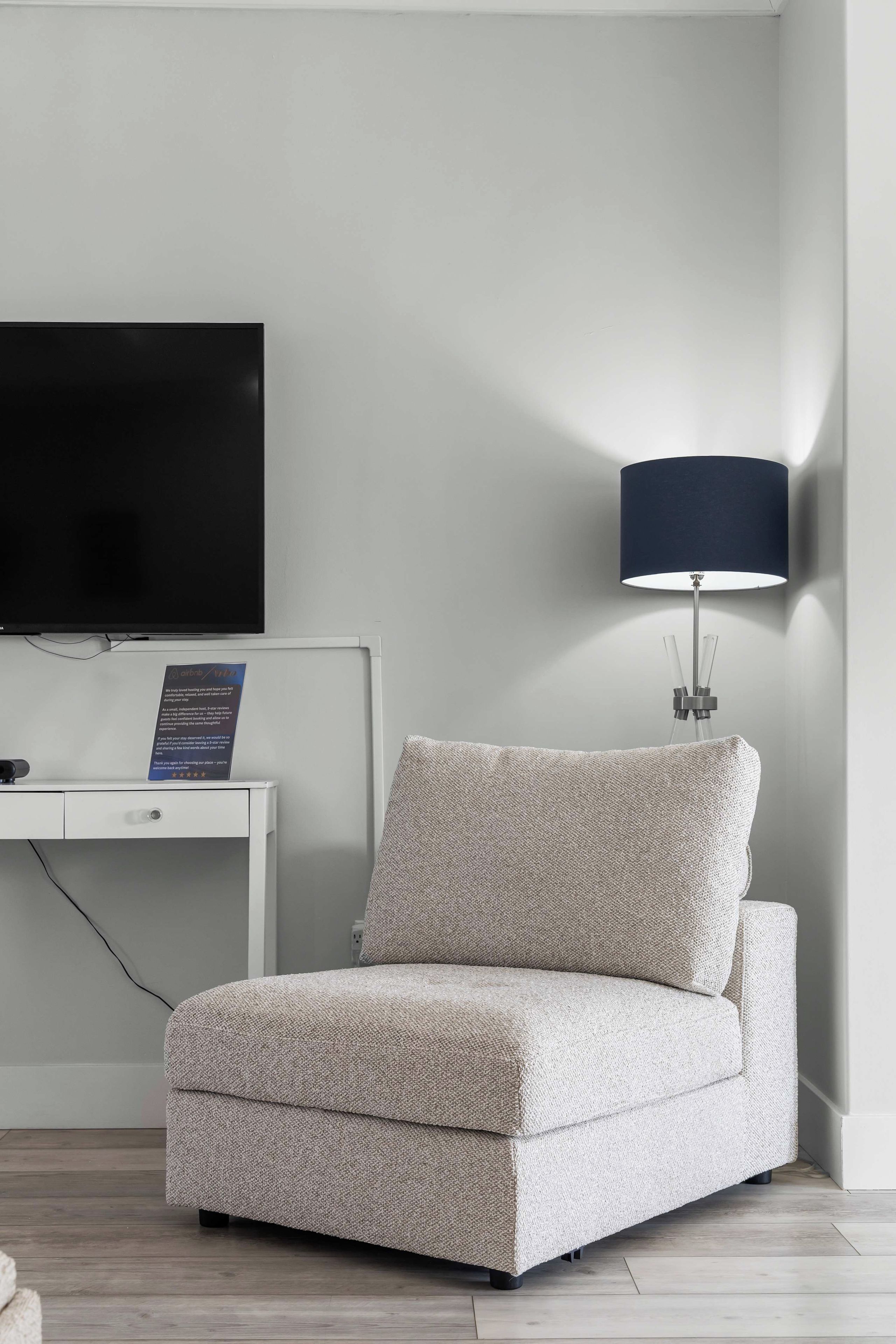 Une chambre d’hôtel moderne avec un téléviseur à écran plat fixé sur une console blanche, un canapé beige et une lampe sur pied.