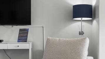 Une chambre d’hôtel moderne avec un téléviseur à écran plat fixé sur une console blanche, un canapé beige et une lampe sur pied.