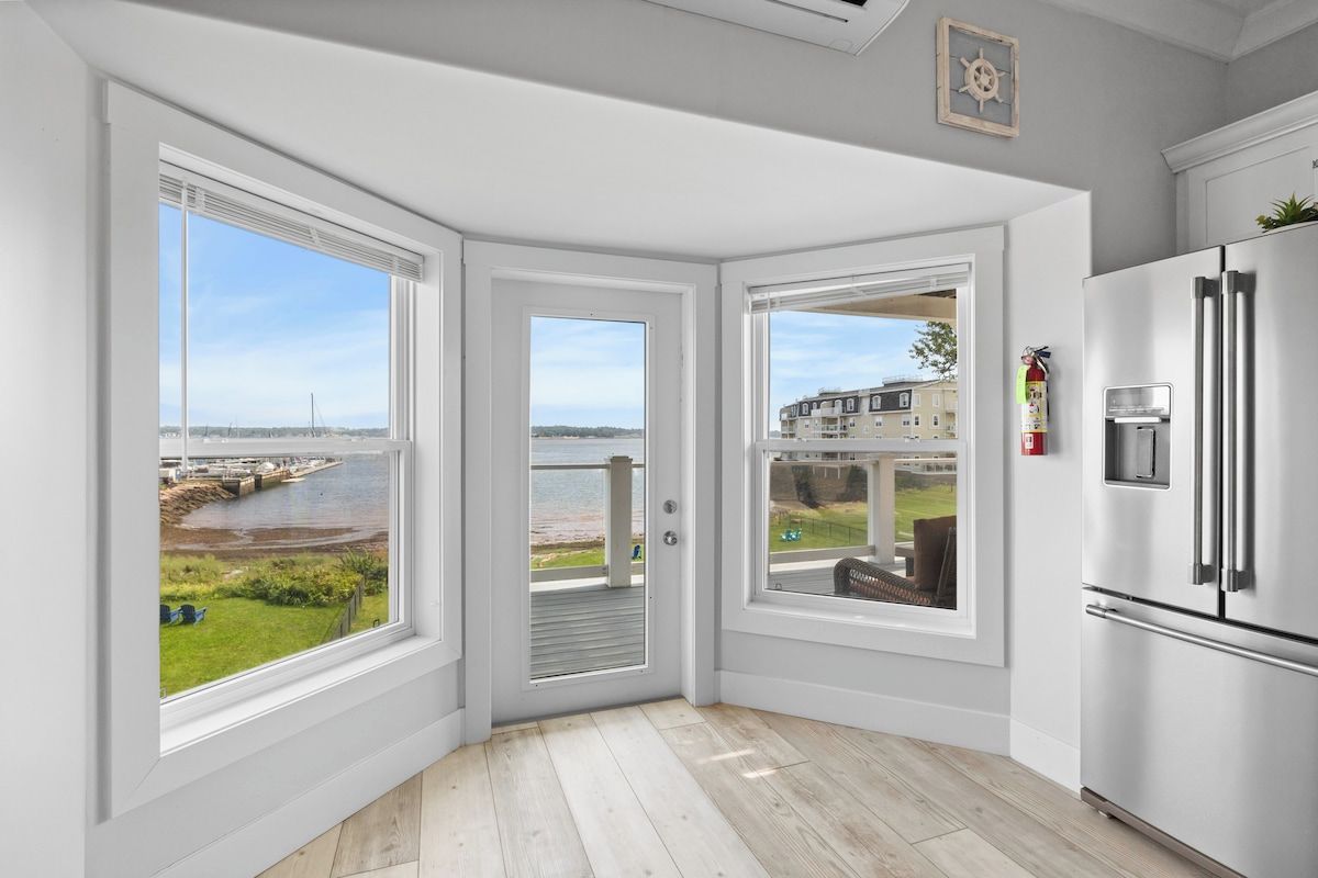 Una cocina con un refrigerador blanco grande, tres ventanas con vistas al frente marítimo y edificios, y piso de madera.