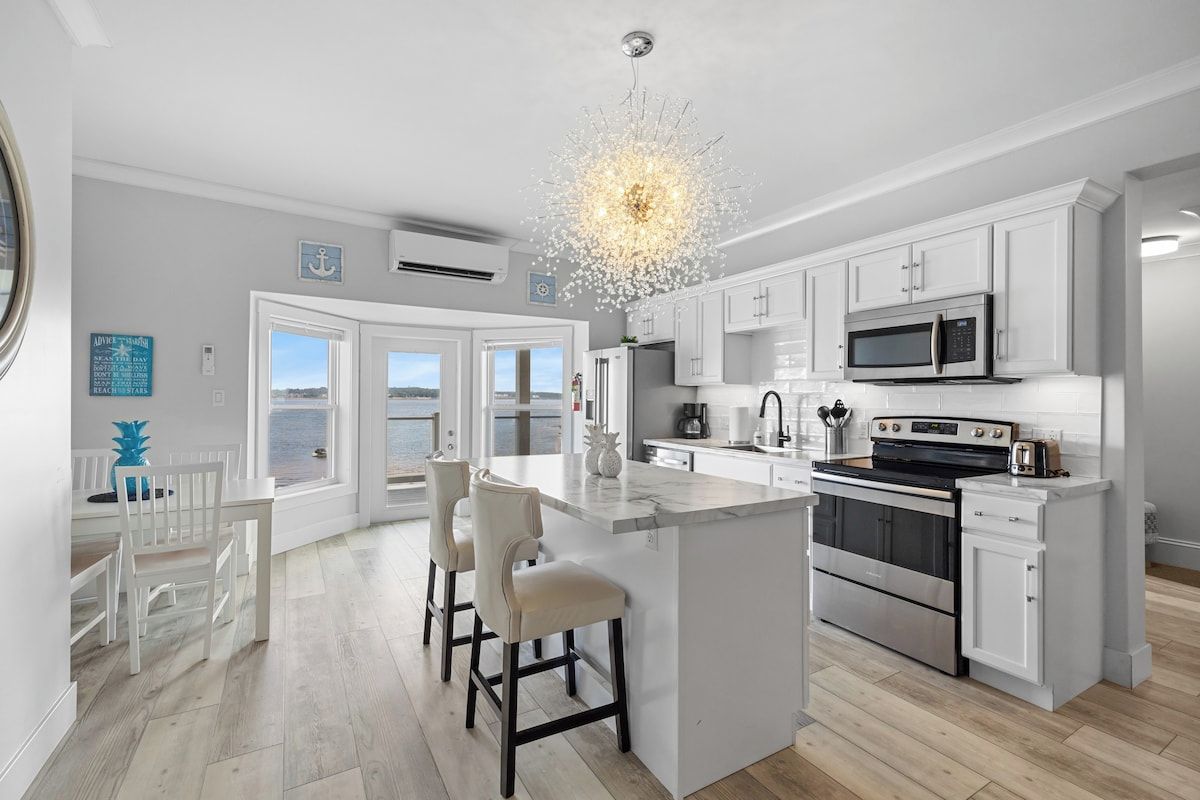 Una cocina moderna con armarios blancos, una isla central con taburetes de bar, una estufa grande en la isla y vistas a través de puertas de vidrio.