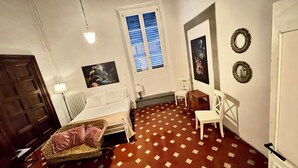 Ein Zimmer mit Schachbrettmuster-Boden, einem Bett, einem Sessel, einem Tisch, einer Lampe, einem Fenster mit Jalousien und Wanddekorationen.