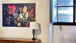 Ein Zimmer mit floralem Gemälde an der Wand, einer Lampe auf einem Holztisch und einem Fenster mit Blick auf ein Gebäude.