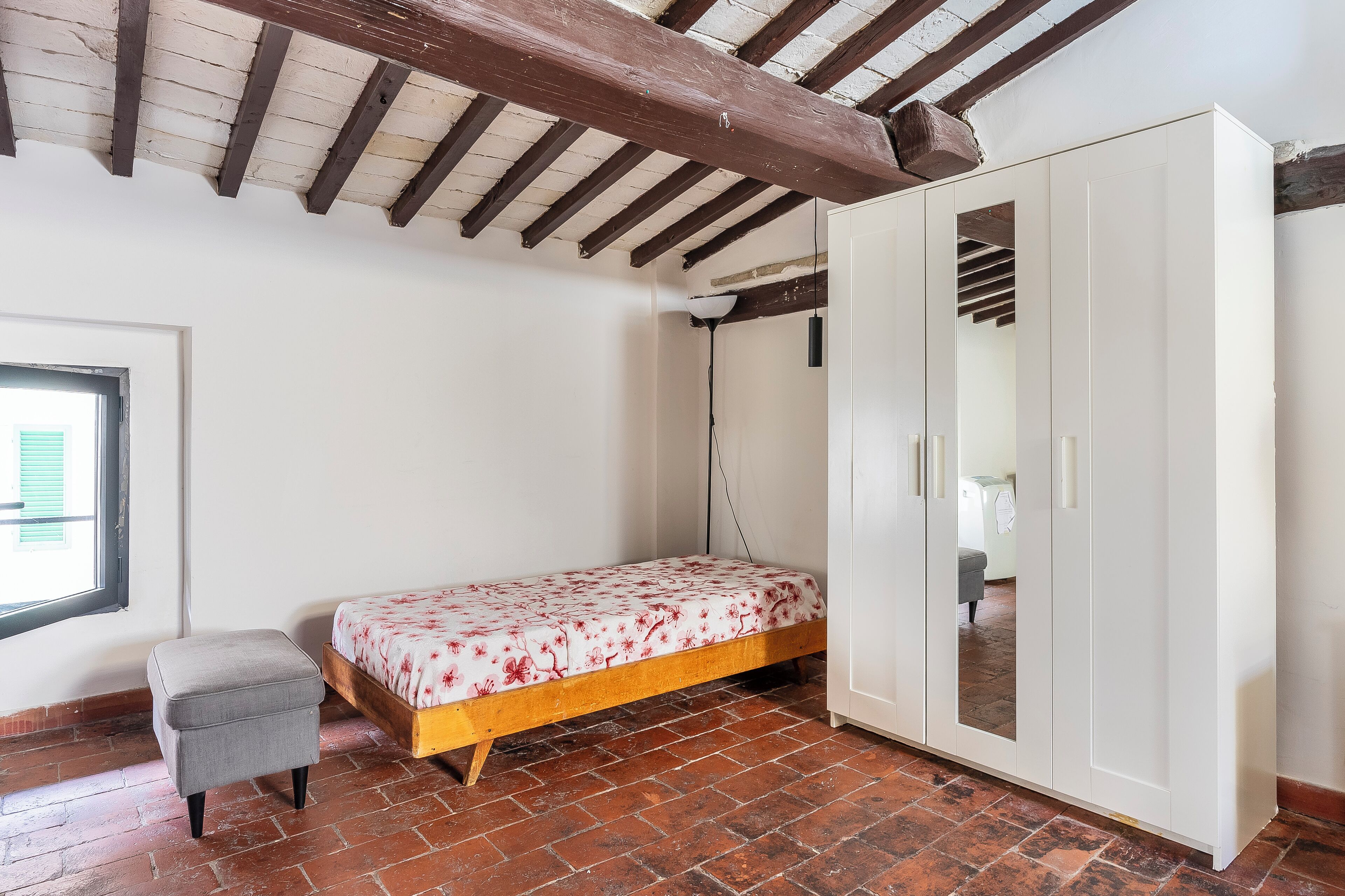 A bedroom with a bed, a small stool, a wardrobe, and a window.