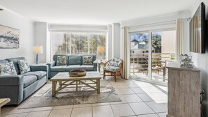 A living room with a sofa, a coffee table, and a chair. There is a window with blinds, a television, and a cabinet.