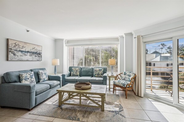A living room with a grey sofa set, a wooden coffee table, and a wicker basket. There is a window with blinds, a lamp, and a chair with a patterned cushion.
