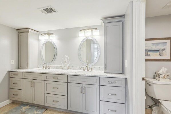 A bathroom with a double vanity, two large oval mirrors, a toilet, and a framed picture on the wall.