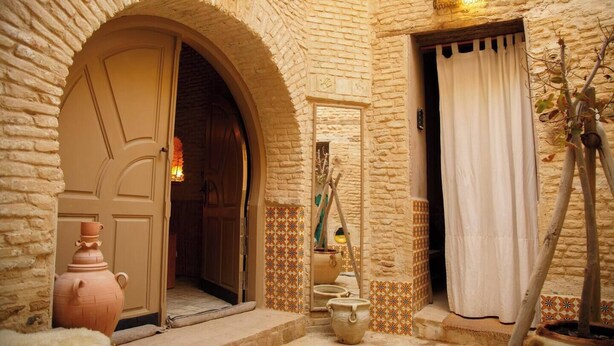 Uma entrada em arco com porta, um pequeno degrau que leva a um espaço revestido de azulejos e uma parede com azulejos decorativos.