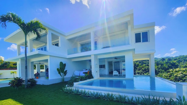 Une maison moderne à deux étages, dotée d’une grande piscine, d’une pelouse verdoyante et luxuriante et de plantes tropicales.