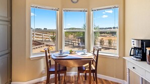 A dining area with a table set for two, three large windows offering a view of a fenced outdoor area, and a coffee maker on a white cabinet. - River Road Retreat! Hot Tub +  Barndominium (Templeton)