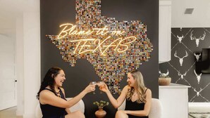Dos personas brindando con bebidas en una habitación que tiene un adorno de pared con el contorno de un estado hecho de botellas y un letrero de neón que dice 'Blame it on Texas'.