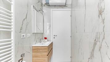 Un bagno moderno con un mobile lavabo bianco, pareti in marmo, un appendi asciugamani, un distributore di asciugamani e un soffione.