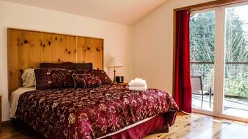 A bedroom with a wooden headboard, a large bed with a red and gold patterned bedspread, a white lamp, and a view of trees through a sliding glass door.