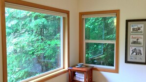 Une pièce d’angle dotée de deux grandes fenêtres donnant sur une forêt. Une petite table en bois, sur laquelle sont posés un bol et un livre, est placée contre le mur.