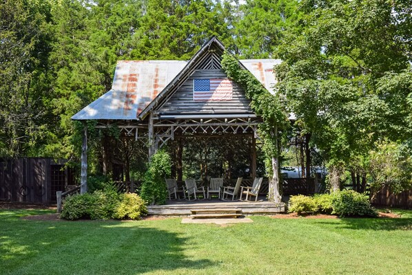 A rustic wooden gazebo with a metal roof, surrounded by lush greenery and a well-maintained lawn.