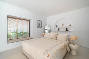 A bedroom with a large bed, a window with blinds, a bedside table with a lamp, a mirror, and a shelf with decorative items. - Villa Janoor by GORO (Seminyak)