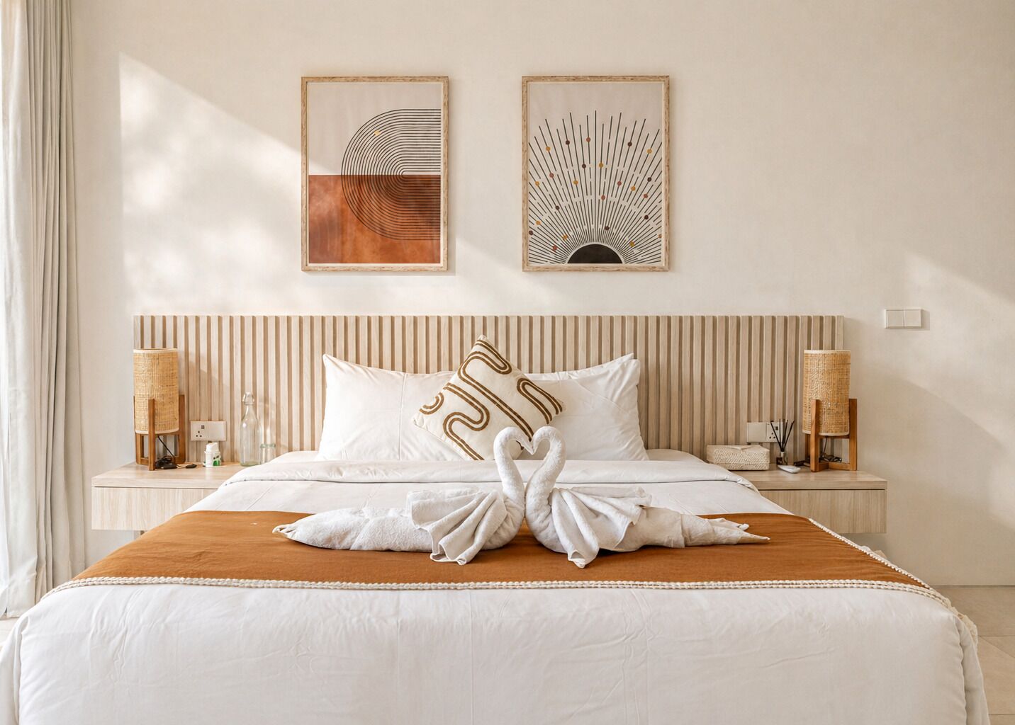 Une chambre d’hôtel avec un lit, deux œuvres d’art accrochées au mur, une tête de lit à rayures, des lampes de chevet et une chaise en osier.