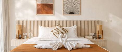 Une chambre d’hôtel avec un lit, deux œuvres d’art accrochées au mur, une tête de lit à rayures, des lampes de chevet et une chaise en osier.
