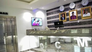 A hotel reception area with a marble counter, a television screen, framed portraits, and clocks on the wall. - DCM Hotel and Suites (Lagos)