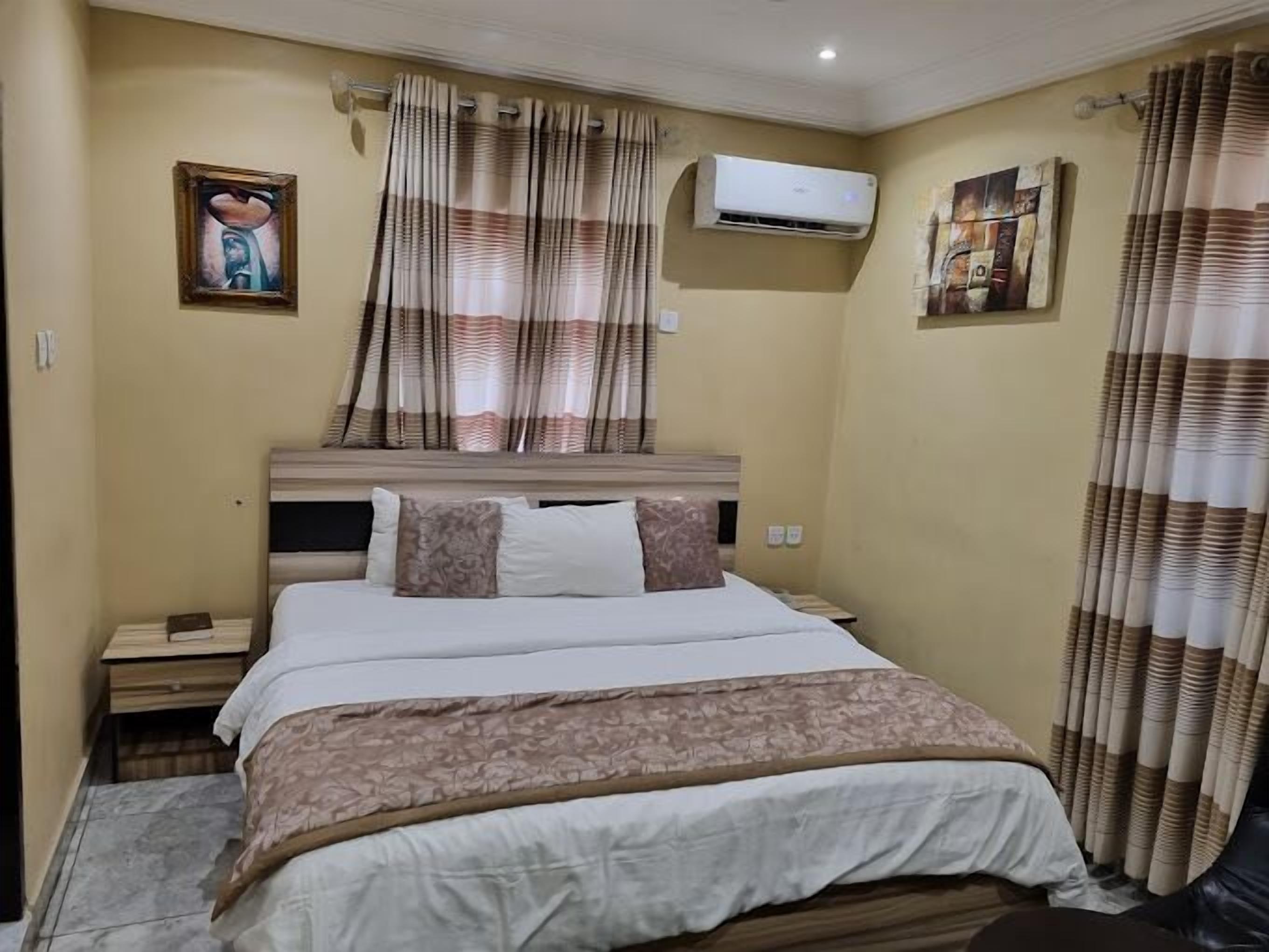 A hotel room with a bed, a nightstand, an air conditioning unit, and curtains.