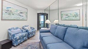 Un salon moderne avec un canapé bleu, des fauteuils assortis et un grand miroir qui reflète des œuvres d’art. On aperçoit au fond une porte sur laquelle est inscrit « MAISON DE PLAGE ».