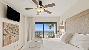 A hotel room with a large bed, a ceiling fan, a TV, artwork, a balcony with a view of the beach and palm trees.