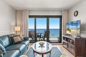 A living room with a sofa, a coffee table, a TV, a balcony with a view of the sea, and a dining area with chairs and a table. - 609 | Chic Beachfront Retreat—1BR, Gulf-View Balcony & VIP Beach Service (Panama City Beach)