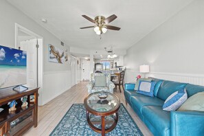 A living room with a blue sofa, a glass coffee table, a ceiling fan, and a painting of a beach scene. - 609 – Beachfront 1BR Escape | Balcony, Gulf Views & VIP Beach Service (Panama City Beach)